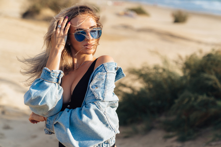 Portrait of a sexy girl with a beautiful figure in a black swimsuit on the beach with sand. Incredible model with blue glasses on her face and a jacket in her hands posingの写真素材