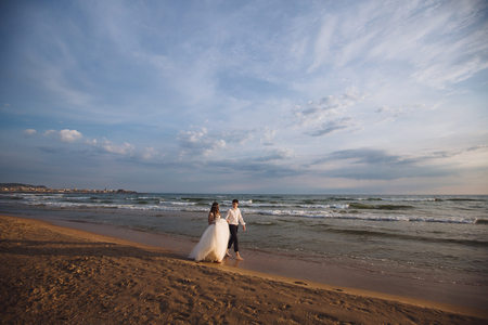 A beautiful couple of newlyweds, the bride and groom walking on the beach. Gorgeous sunset and sky. Wedding dresses, a white luxury dress for a girl. Family concept, honeymoon.の写真素材