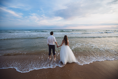 A beautiful couple of newlyweds, the bride and groom walking on the beach. Gorgeous sunset and sky. Wedding dresses, a white luxury dress for a girl. Family concept, honeymoon.の写真素材