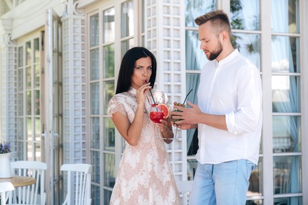 A fragile beautiful young brunette in a lacy beige dress drinks her delicious berry cocktail through a straw, stands next to her brutal tall guy with a beard. Lovers celebrate something.の写真素材