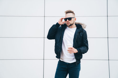 Stylish young man in sports jacket, sunglasses and coffee in his hands standing on background of...