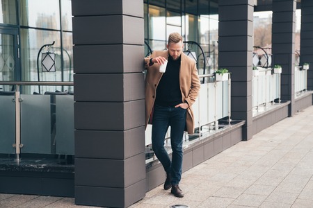Attractive man in trendy brown coat and coffe in his hand posing near business centerの写真素材