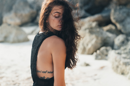 Portrait of sexy brunette model in knitted sweater posing on the sandy beach, big stones and rock on background.の写真素材