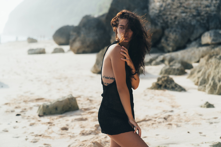 Portrait of sexy brunette model in knitted sweater posing on the sandy beach, big stones and rock on background.の写真素材