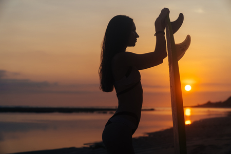 Silhouette of slim girl with surfboard in hands at beach on background of beautiful sunsetの写真素材
