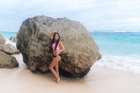 Sexy tanned girl in swimsuit standing on beach background of large stones and oceanの写真素材