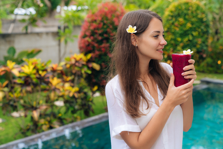 Beautiful brunette with flower in her hair and glasses of fresh juice stands in tropical garden.の写真素材
