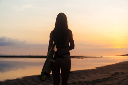 Backside view silhouette of surfer girl with surf board on beach at sunsetの写真素材