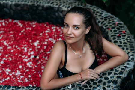 Beautiful smiling girl relaxing in bath with rose petals and tropical flowers outdoor at spa dayの写真素材