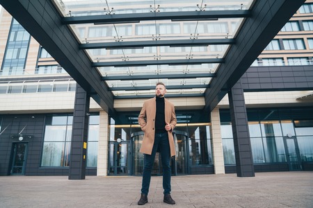 Attractive man in trendy brown coat posing near business center. Handsome european man with beard looks away. Concept lifestyle, fashionable men styleの写真素材