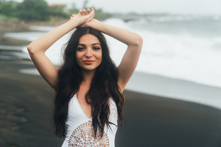Portrait of beautiful girl in white swimsuit posing with raised arms on beach with black sandの写真素材