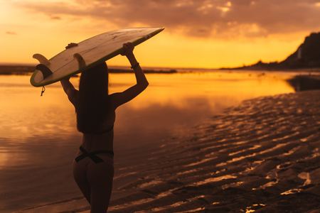 Silhouette of slim girl with surfboard in hands at beach on background of beautiful sunsetの写真素材