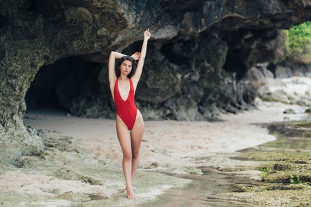 Attractive slender girl in red swimwear posing with rised arms near rock on beachの写真素材