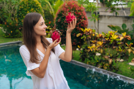 Happy girl with beautiful smile holds two dragon fruits, pitaya in her hands background of poolの写真素材