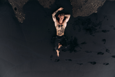Sexy muscular guy in pants lies on black volcanic beach near big stone.の写真素材