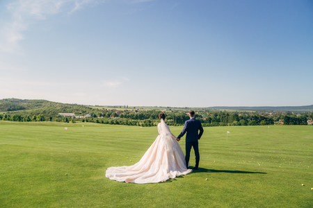 Backside view newlyweds couple holds hands each other, walks on golf club gladeの写真素材