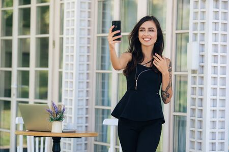 Beautiful girl in dress with tattoo on her hand takes selfie on phone in cafeの写真素材