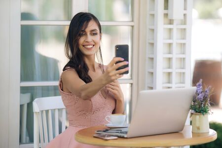 Beautiful girl in dress with tattoo on her hand takes selfie on phone in cafeの写真素材