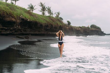 Beautiful girl in dark swimsuit walks on black sand beach near oceanの写真素材