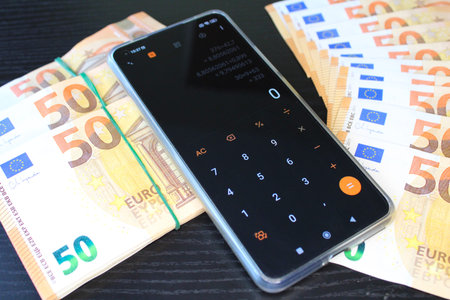 Several stacks of fifty euro banknotes and a smartphone with a running calculator lie on a black...
