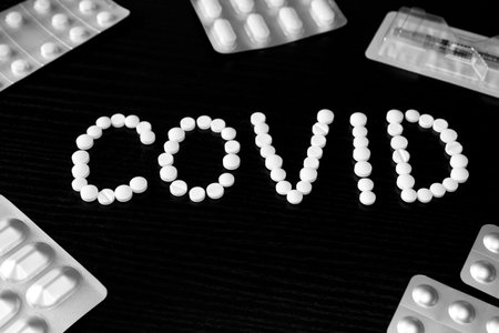 The word COVID, laid out from white tablets, on a black background, surrounded by tablets and a...