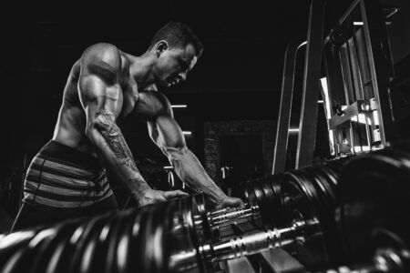 Muscular athletic bodybuilder man hard workout in gym over dark background with dramatic light with barbellの写真素材