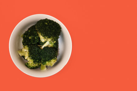 steamed green broccoli in a white bowl on an orange-carrot color background, top view boiled...