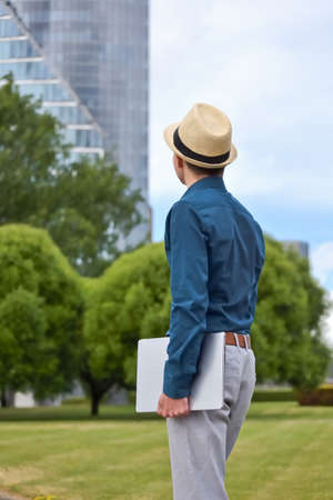 A man with a laptop in his hand stands and looks towards the office building. Young student, programmer and businessman with computer in hand looks at office building and thinks about work and career.の写真素材