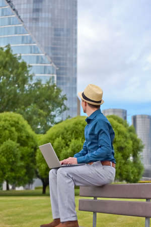 A man with a laptop works remotely on a bench outdoors and looks at an office building.の写真素材