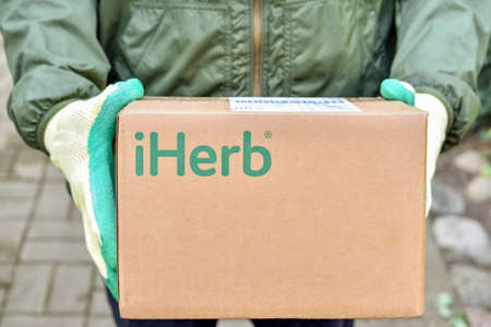 Logotype iHerb on cardboard box in courier hands. iHerb is an herbal products company, provides and delivers health and wellness products to people around the world: Riga, Latvia - September 10, 2021のeditorial素材