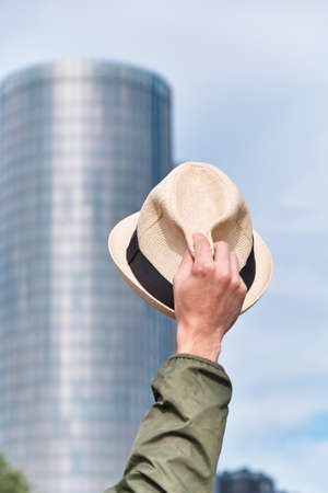 Man's hat in hand against the backdrop of a skyscraper office building and blue sky. Welcome and greeting hand gesture, goodbye office and freedom concept.の写真素材