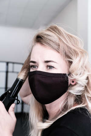 Blonde woman in a protective mask curls her hair with a curling iron.の写真素材