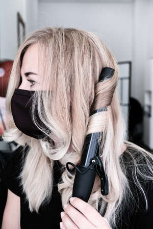 Woman in a protective mask curls her blond hair with a curling iron.の写真素材