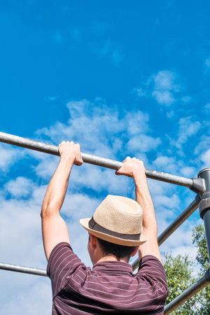Back view of a young man pull-up on the bar against the sky, copy space. Hipster or nerd exercising...