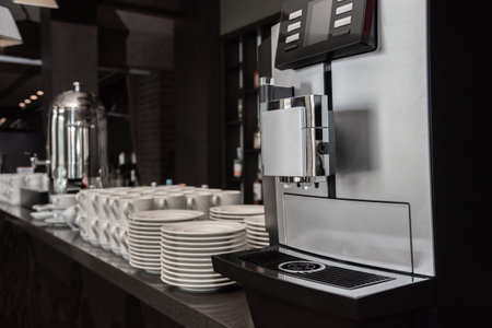 Automatic coffee machine against the background of blurred interior with mugs and plates on the bar prepared for self-service breakfast buffet.の写真素材