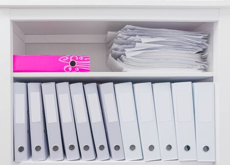 White office folders standing on a shelf next to a pink folder and a stack of papers.の写真素材