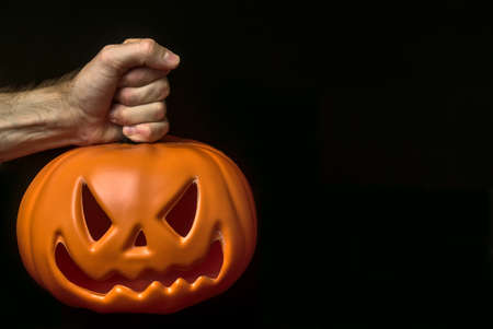 Man hand holding onto pumpkin stem. Scary Halloween 2020 theme. Beautiful male hand keeping isolated glowing pumpkin on black background with copy spaceの写真素材
