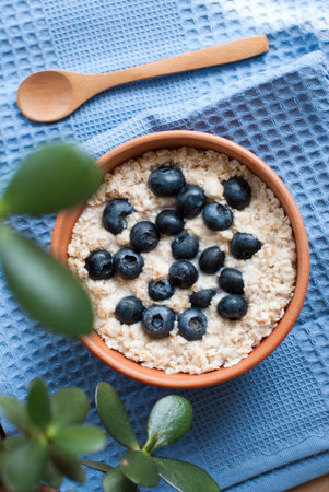 Oatmeal porridge with blueberries and wooden spoon on a blue towel, tasty healthy summer breakfast
