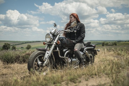 The red-haired biker girl is sitting on a motorcycle. field of meadow and cloudsの写真素材