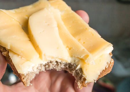 Bitten sandwich made of rye bread, butter and cheese in a man's hand in the rays of a bright morning sun close.の写真素材