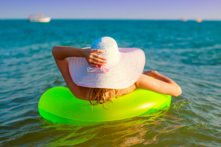 Girl in a hat with wide fields floats on a sea wave on a bright green inflatable round mattress rear view.の写真素材