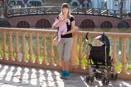 Mom with a baby girl and a stroller on the background of a pond and a bridgeの写真素材