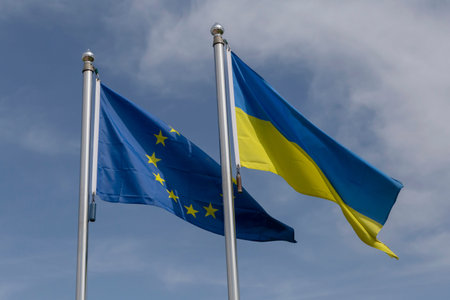 European Union and Ukrainian flags on flagpoles against blue sky with white clouds