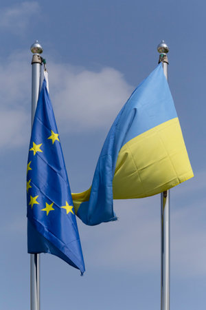 European Union and Ukrainian flags hanging on flagpoles against blue sky with cloudsの写真素材