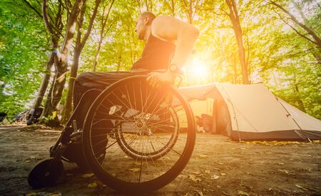 Disabled man resting in a campsite with friends. Wheelchair in the forest on the background of tents. Camping. Summertimeの写真素材