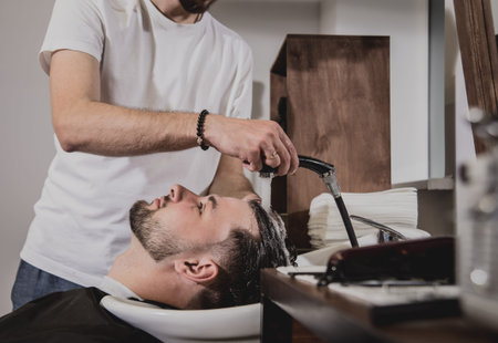 Young man with trendy haircut at barber shop. Barber washes customer head.