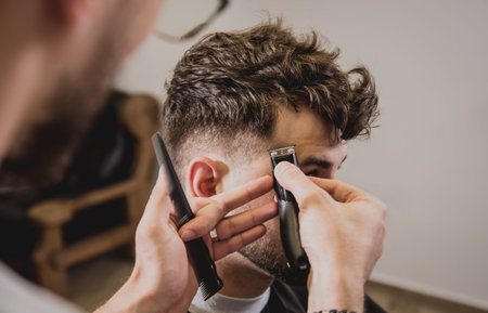 Young man with trendy haircut at barber shop. Barber does the hairstyle and beard trim. Concept...