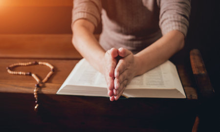 Christian woman praying in church. Hands crossed and Holy Bible on wooden desk.