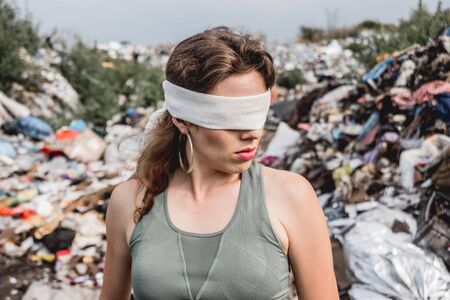 Blindfolded female volunteer in a landfill of plastic trash. Bushes and sky in the background. Earth day and ecology. Conceptの写真素材