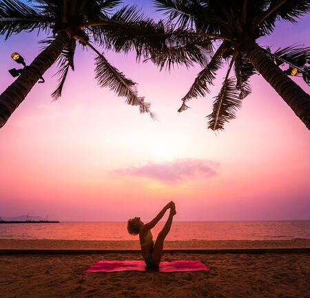 Beautiful young woman practic yoga at the beach. Early morning exercise. Sunrise. Palms backgroundの写真素材